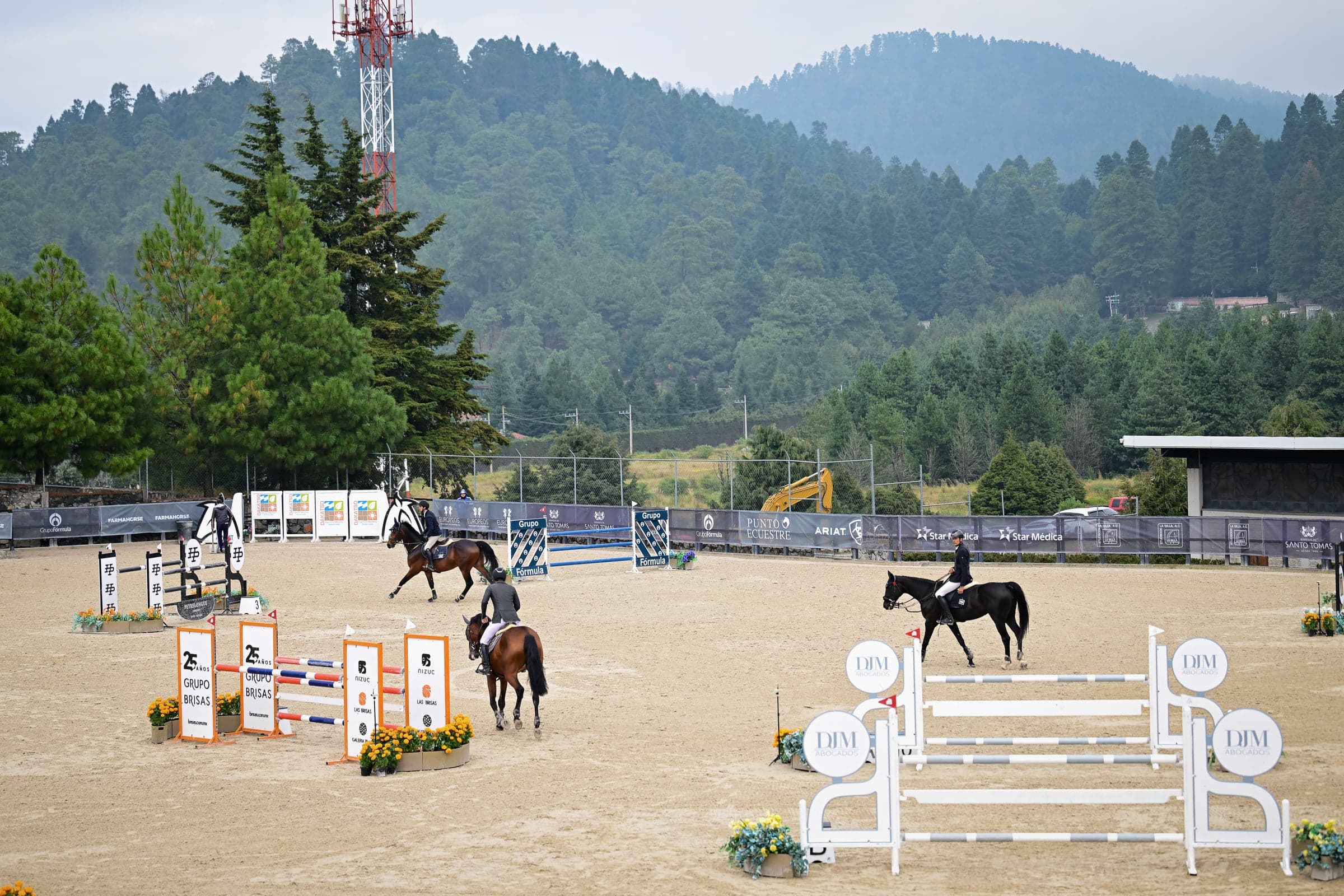 Pista de la Copa con obstáculos de patrocinadores y jinetes a lo lejos, frente a las montañas boscosas.