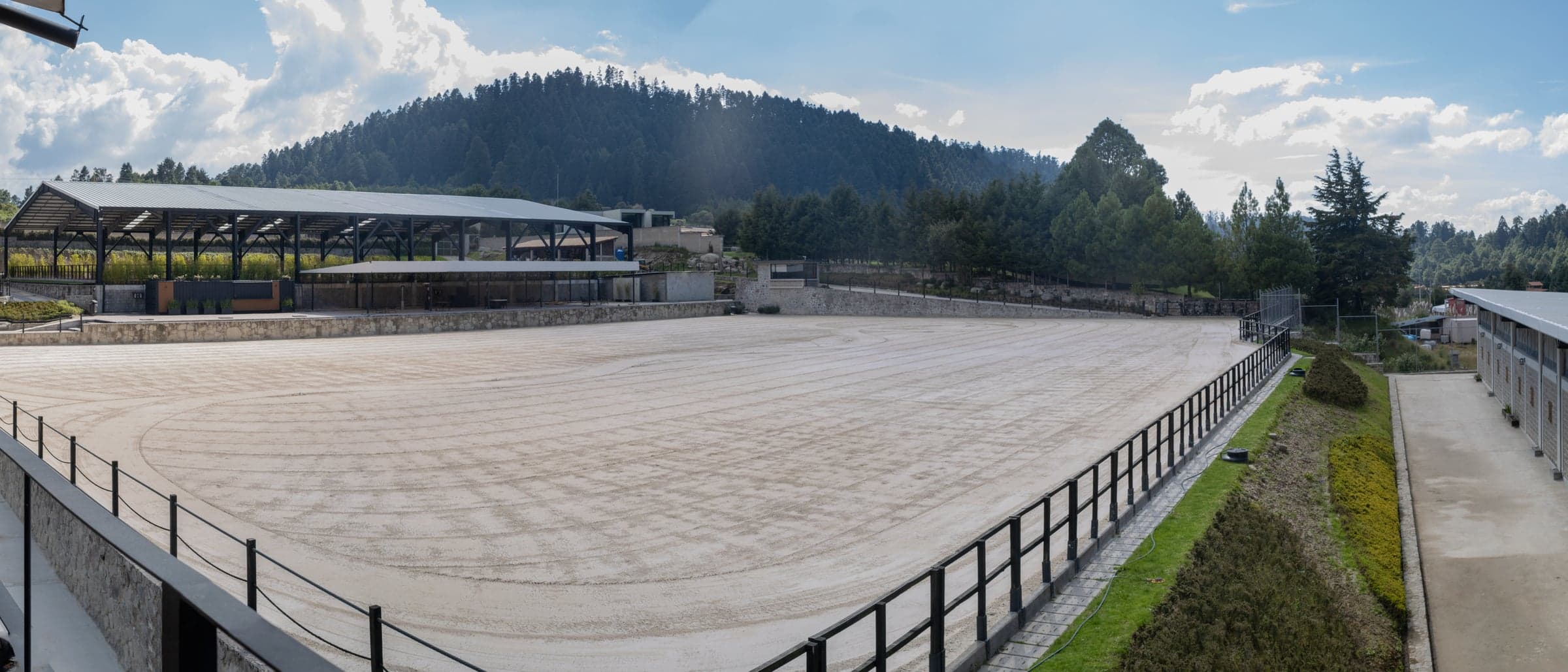 Panorámica de la pista principal de arena rastrillada, con la pista techada a la izquierda y un cerro boscoso al fondo.